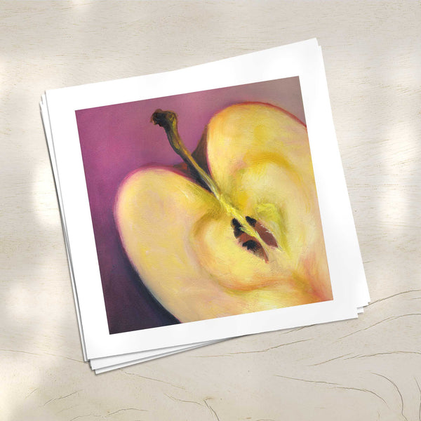A stack of square art prints on archival paper on a wood kitchen counter. The prints show a cream apple cut in half to show the subtle love heart at the core, on a soft pink background. This is a giclee print of my realistic oil painting still life. The original artwork is sold.