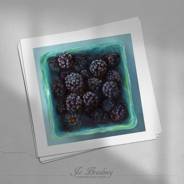 A stack of square art prints on archival paper on a stone kitchen counter. The prints show fresh blackberries in a teal farmers market box. This is a giclee print of my realistic oil painting still life.