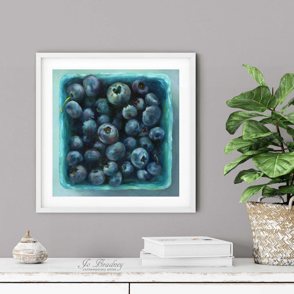 A blueberry oil painting print in a simple elegant white frame on a dining room or living room wall. There are books and a plant in a rustic pot, on a shabby chic painted wood buffet table.