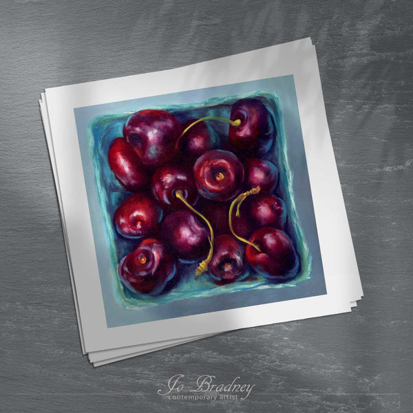 A stack of square art prints on archival paper on a slate kitchen counter. The prints show fresh summer cherries in a farmers market box. This is a giclee print of my realistic oil painting still life.