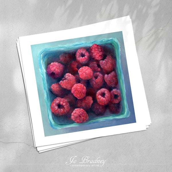 A stack of square art prints on archival paper on a stone kitchen counter. The prints show fresh summer raspberries in a farmers market box. This is a giclee print of my realistic oil painting still life. The original artwork is sold.