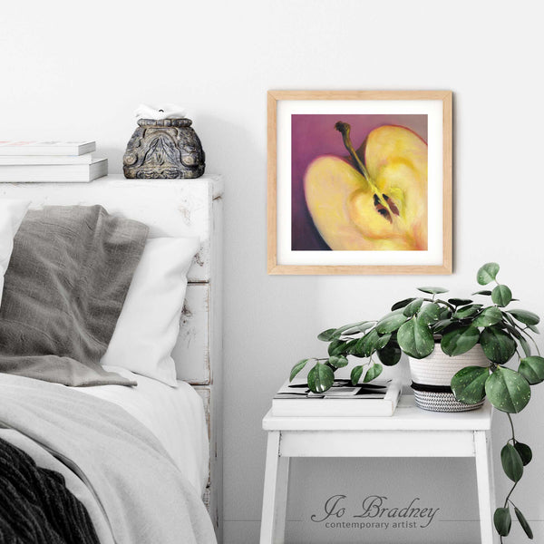 A subtle love heart art print of an apple oil painting in a simple, elegant wood picture frame on a bedroom wall with a shabby chic bedside table, potted plant and books. The smallest square print is 4x4, the largest is 12x12 inches.