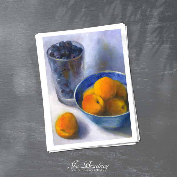 A stack of vertical art prints on archival paper on a slate kitchen counter. The prints show blueberries in a pint glass and orange apricots in a blue china bowl. This is a giclee print of my realistic oil painting fruit still life. The original artwork is sold.