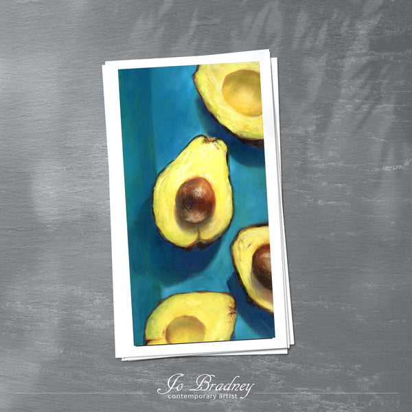A stack of tall narrow art prints on archival paper on a slate kitchen counter. The prints show creamy yellow avocados on a deep turquoise background. This is a giclee print of my realistic oil painting still life. 