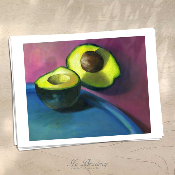 A stack of horizontal art prints on archival paper on a wood kitchen counter. The prints shows a green avocado on purpleand teal background. This is a giclee print of my realistic oil painting vegetable still life. 