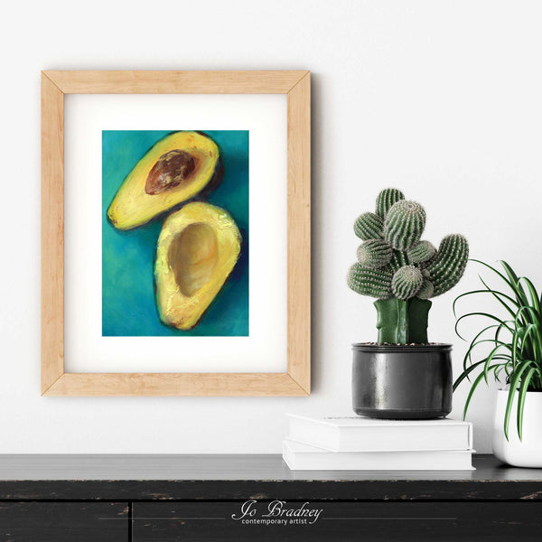 A fresh avocado painting art print in a simple, elegant wood picture frame on a living room or dining room wall, with a pile of books and a cactus. The print size ranges from 4x6 to 11x14 inches.