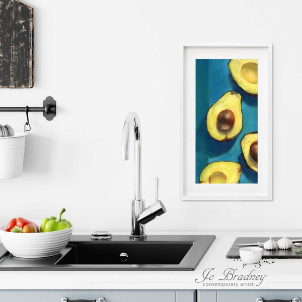 A tall narrow avocado painting art print in a simple, elegant white picture frame on a kitchen wall. Placed above a modern sink, with rustic chopping boards and fresh vegetables.