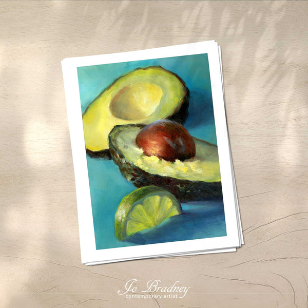 A stack of vertical art prints on archival paper on a wood kitchen counter. The prints show fresh avoacdo and slice of lime on a teal background. This is a giclee print of my realistic oil painting still life.