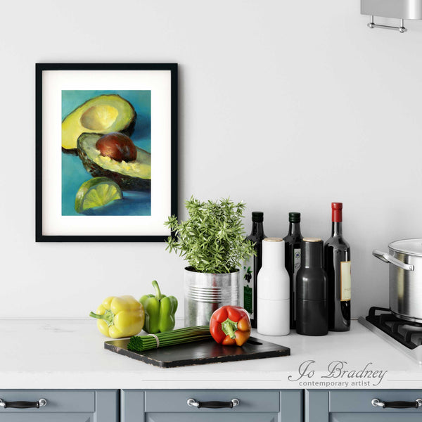 A bright turquoise avoaacdo painting print in a simple, elegant black picture frame on a kitchen wall. There is a chopping board, vegetables, herbs and some bottles. The art print is as large as three bell peppers. The largest size is 14 inches high by 11 wide