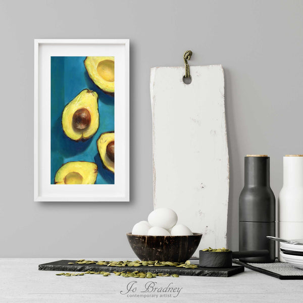 A narrow avocado art print in a simple, elegant white picture frame on a kitchen wall. There is a bowl of eggs, and a chopping board for scale. The print is 5 eggs tall. The smallest skinny art print is 4x8 inches, the largest is 10x20 inches.