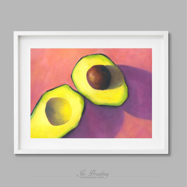 A yellow and green avocado cut in half on a bright pink background. This archival art print of my vegetable still life oil painting is shown in simple white picture frame.