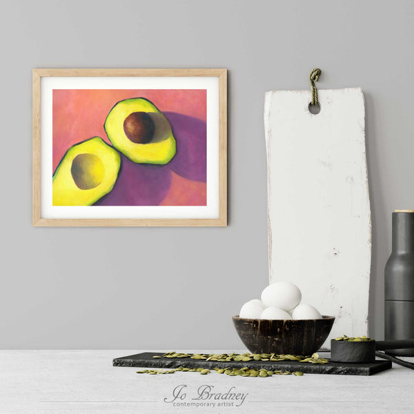 A pinkand green avocado art print in a simple, elegant wood picture frame on a kitchen wall. There is a bowl of eggs, and a chopping board for scale. The print is 5 eggs tall. The smallest horizontal print is 4x6 inches, the largest is 11x14 inches.