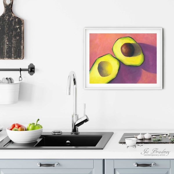 A horizontal avocado painting art print in a simple, elegant white picture frame on a kitchen wall. Placed above a modern sink, with rustic chopping boards and fresh vegetables.