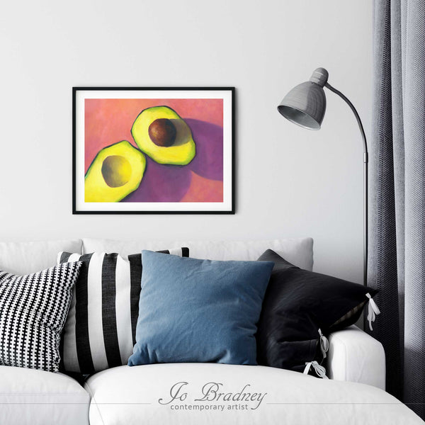 A bright avoacdo print in a simple, elegant black frame with a pile of sofa cushions on a college dorm, game room or living room wall. The art print is the same size as a cushion. The largest size is 11x14 inches , the smallest is 4x6 inches.