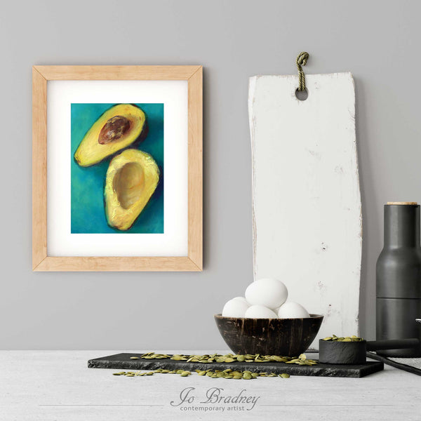 An avocado art print in a simple, elegant wood picture frame on a kitchen wall. There is a bowl of eggs, and a chopping board for scale. The print is 5 eggs tall. The smallest vertical print is 4x6 inches, the largest is 11x14 inches.
