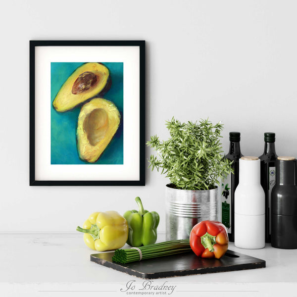 An avocado painting giclee fine art print in a simple, elegant black picture frame on a kitchen wall. There is a chopping board, vegetables, herbs and some bottles. The art print is as large as three bell peppers. The largest size is 11 inches high by 14 wide