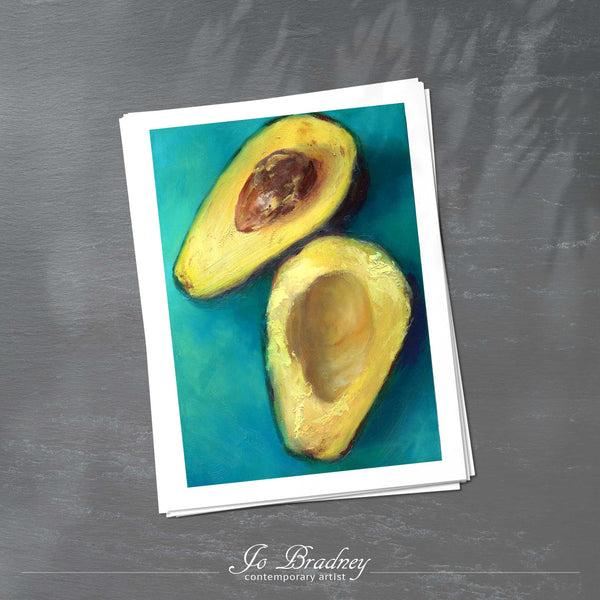 A stack of vertical art prints on archival paper on a slate kitchen counter. The prints show a fresh avocado cut in half and twisted on a dark turquoise background. This is a giclee print of my realistic oil painting still life.