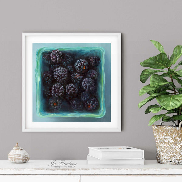 A blackberry oil painting print in a simple elegant white frame on a dining room or living room wall. There are books and a plant in a rustic pot, on a shabby chic painted wood buffet table.