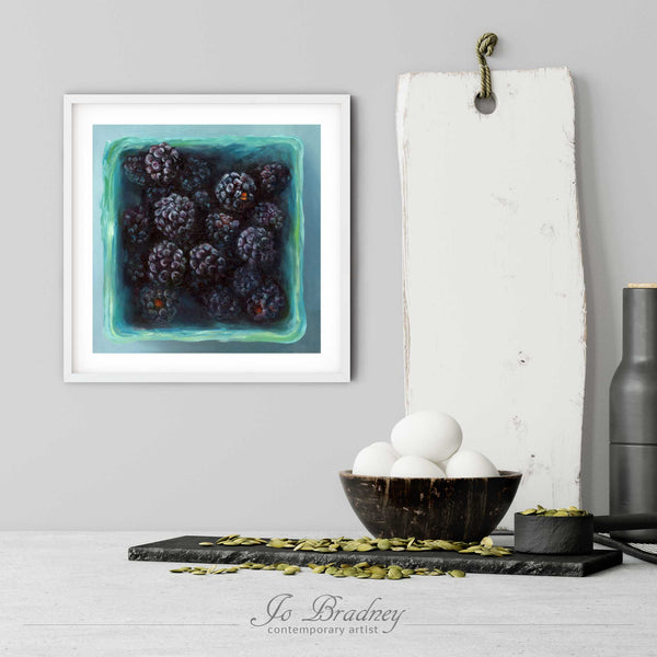 A blackberry art print in a simple, elegant white picture frame on a kitchen wall. There is a bowl of eggs, and a chopping board for scale. The print is 5 eggs tall. The smallest square print is 4 inches, the largest is 12 inches.