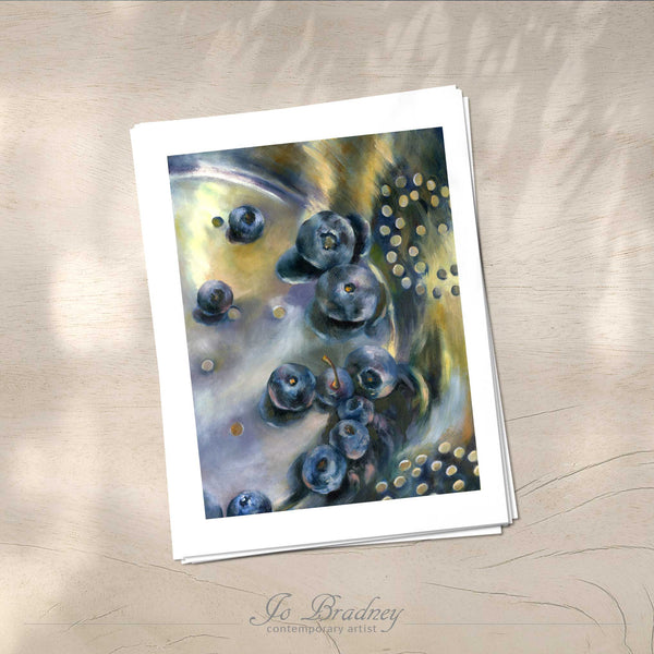 A stack of vertical art prints on archival paper on a wood kitchen counter. The prints show fresh wash blueberries in a silver stainer. This is a giclee print of my realistic oil painting still life. The original artwork is sold.