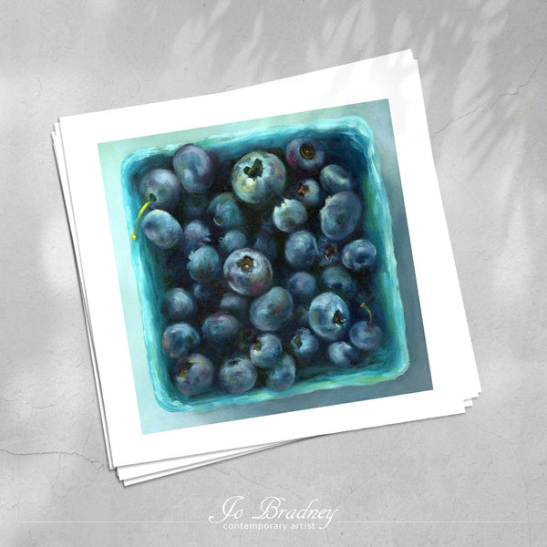 A stack of square art prints on archival paper on a stone kitchen counter. The prints show fresh summer blueberries in a teal market box. This is a giclee print of my realistic oil painting still life.