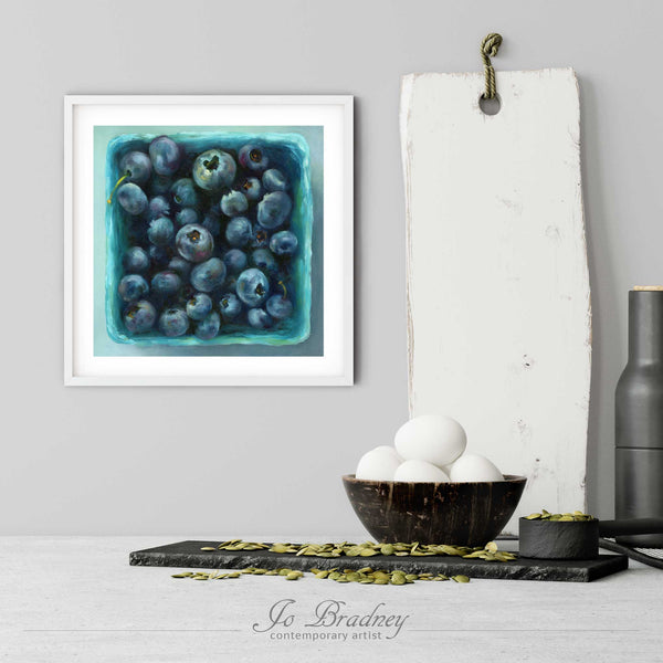 A blueberry art print in a simple, elegant white picture frame on a kitchen wall. There is a bowl of eggs, and a chopping board for scale. The print is 5 eggs tall. The smallest square print is 4 inches, the largest is 12 inches.