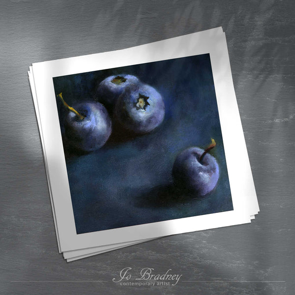 A stack of square art prints on archival paper on a slate kitchen counter. The prints show a dark indigo blue background with four plump blueberries. This is a giclee print of my realistic oil painting still life. The original artwork is sold.