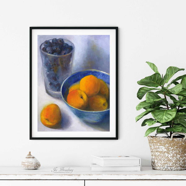 A bowl of apricots and a pint glass of blueberries oil painting art print in a simple elegant black frame on a dining room or living room wall. There are books and a plant in a rustic pot, on a shabby chic painted wood buffet table.