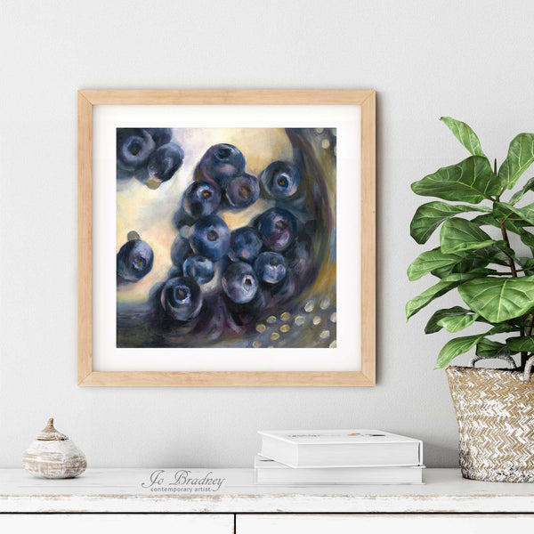 A blueberry oil painting art print in a simple elegant wood frame on a dining room or living room wall. There are books and a plant in a rustic pot, on a shabby chic painted wood buffet table.