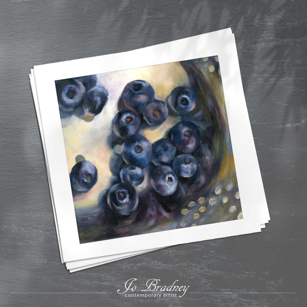 A stack of square art prints on archival paper on a slate kitchen counter. The prints show fresh blueberries washed in a silver colander. This is a giclee print of my realistic oil painting fruit still life. The original artwork is sold.