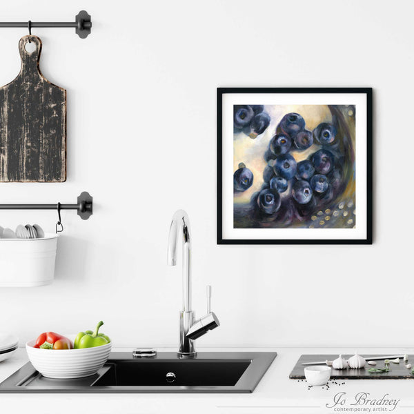 A square art print of washed blueberries in a silver strainer, framed in a simple, elegant black frame on a kitchen wall. Placed above a modern sink, with rustic chopping boards and fresh vegetables.
