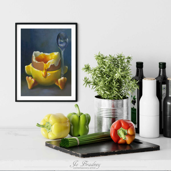 A boiled egg painting art print in a simple, elegant black picture frame on a kitchen wall. There is a chopping board, vegetables, herbs and some bottles. The art print is as large as three bell peppers. The largest size is 14 inches high by 11 wide