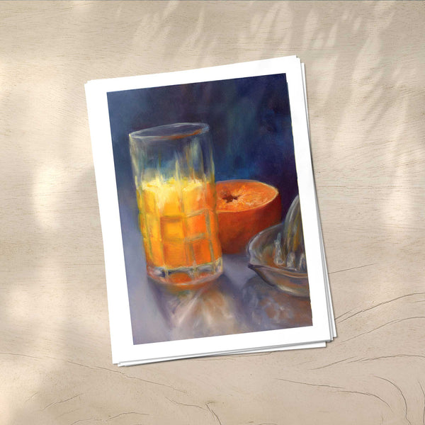 A stack of vertical art prints on archival paper on a wood kitchen counter. The prints show a glass of fresh squeezed orange juice on a dark indigo blue background. This is a giclee print of my realistic oil painting still life. 
