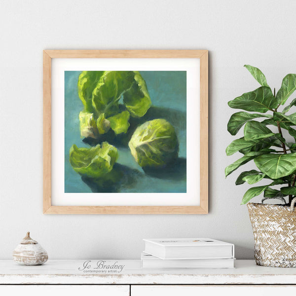 A brussel sprout oil painting art print in a simple elegant wood frame on a dining room or living room wall. There are books and a plant in a rustic pot, on a shabby chic painted wood buffet table.
