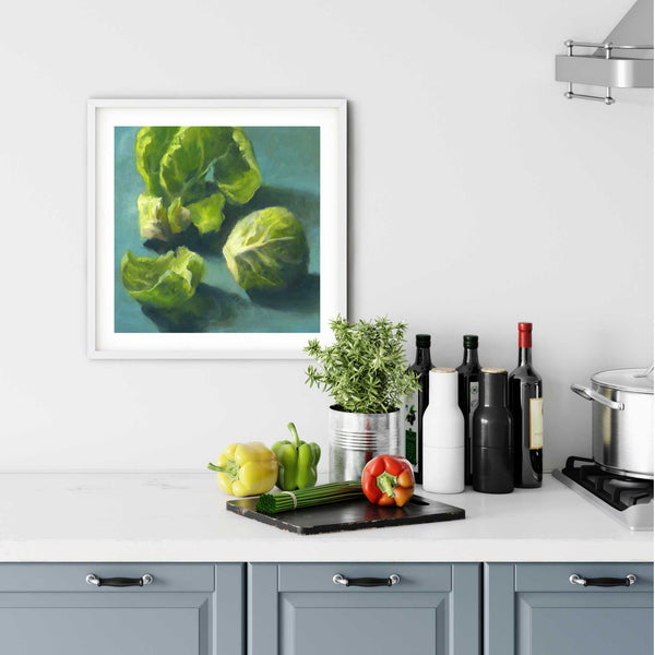 A Brussel Sprouts oil painting print in a simple, elegant white frame on a kitchen wall. There is a chopping board, vegetables, herbs and some bottles. The art print is as large as three bell peppers. The largest size 12 inches square, the smallest is 4 inches.