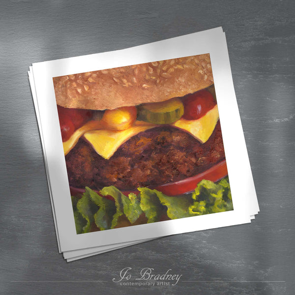 A stack of square art prints on archival paper on a slate kitchen counter. The prints show a close of a cheese burger in a bun. This is a giclee print of my realistic food oil painting still life. The original artwork is sold.
