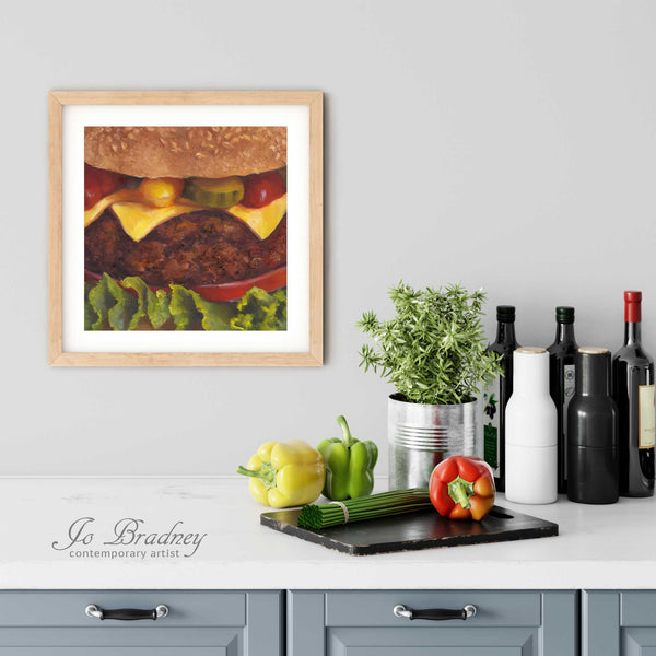 A burger oil painting giclee art print in a simple, elegant wood picture frame on a kitchen wall. There is a chopping board, vegetables, herbs and some bottles. The art print is as large as three bell peppers. The largest size 12 inces square, the smallest is 4 inches.