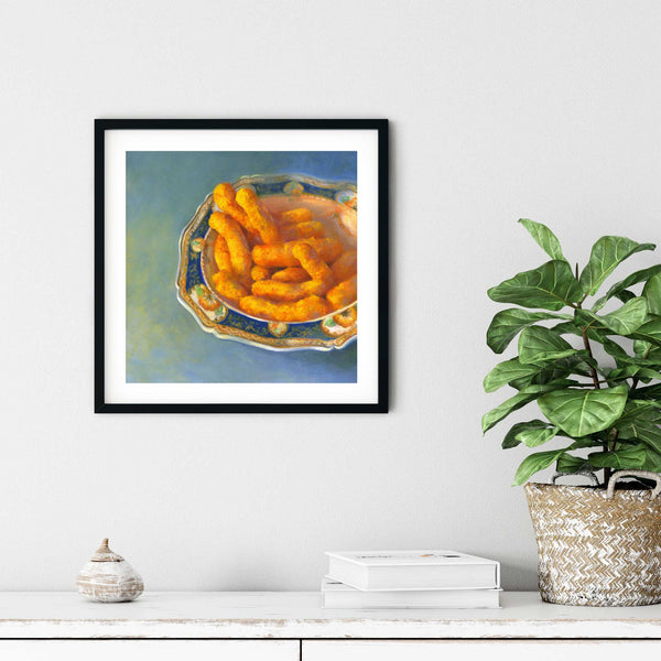 A snack food oil painting art print of cheese puffs in a porcelain bowl in a simple elegant black picture frame on a dining room or living room wall. There are books and a plant in a rustic pot, on a shabby chic painted wood buffet table.