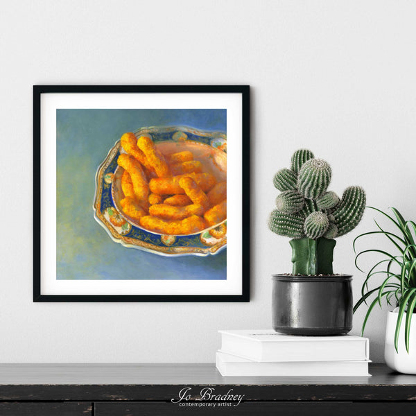 A cheese puff snack food painting art print in a simple, elegant picture frame on a living room or dining room wall, with a pile of books and a cactus. The print size ranges from 4 to 12 inches square.