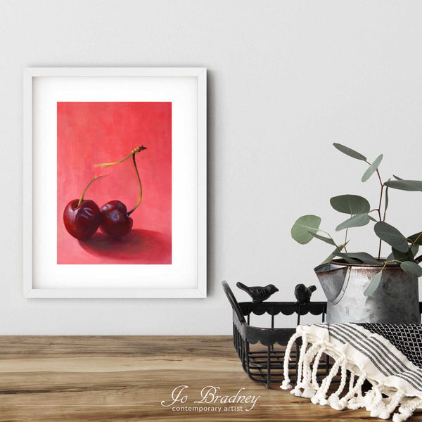 A colorful pink and red cherry art print in a simple, elegant white picture frame on a dining room or boho living room wall. There are flowers in a teapot on a metal tray set on a wood table. The smallest vertical print is 4x6 inches, the largest is 11x14 inches.