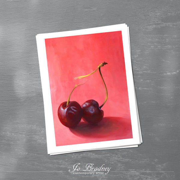 A stack of vertcal art prints on archival paper on a slate kitchen counter. The prints show two red cherries on a deep rose pink background. This is a giclee print of my realistic oil painting still life. 