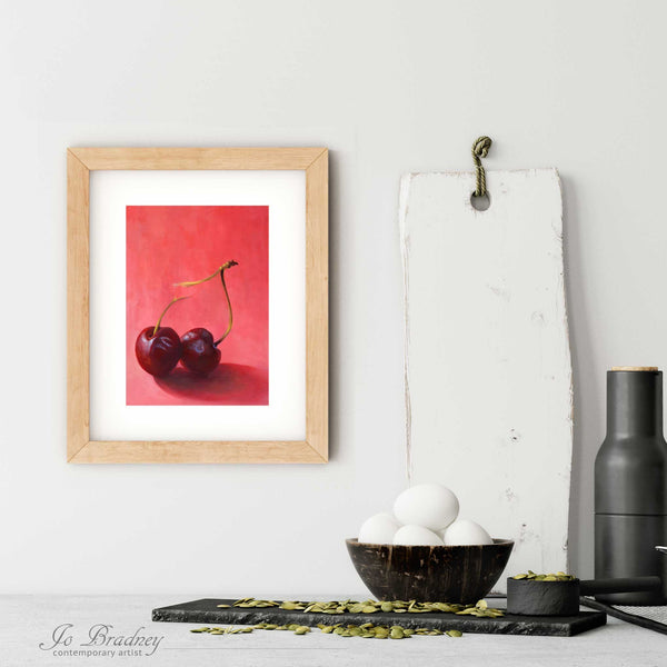 A pink and red cherry art print in a simple, elegant wood picture frame on a kitchen wall. There is a bowl of eggs, and a chopping board for scale. The print is 5 eggs tall. The smallest vertical print is 4x6 inches, the largest is 11x14 inches.
