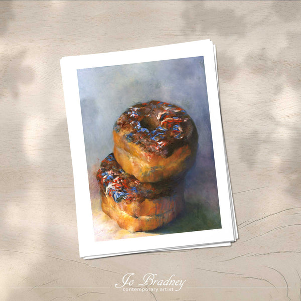 A stack of vertical art prints on archival paper on a wood kitchen counter. The prints show chocolate donuts with sprinkles. This is a giclee print of my realistic oil painting snack food still life. The original artwork is sold.