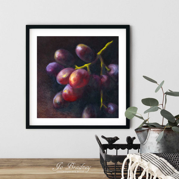 A dark purple and red grapes painting art print in a simple, elegant black picture frame on a dining room or living room wall. There are flowers in a teapot on a metal tray set on a wood table. The smallest square print is 4 inches, the largest is 12 inches.