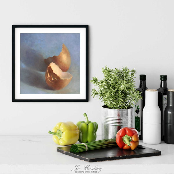 A blue and brown egg oil painting art print in a simple, elegant black frame on a kitchen wall. There is a chopping board, vegetables, herbs and some bottles. The art print is as large as three bell peppers. The largest size is 12 inches square, the smallest is 4 inches.