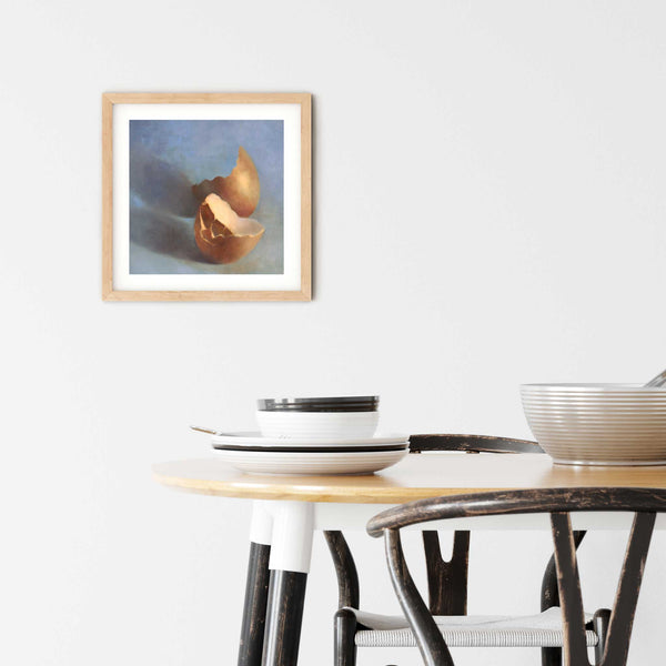 A blue and brown egg art print of my oil painting on a kitchen wall. It is framed in an elegant wood frame and is shown with a dining table laid for breakfast.