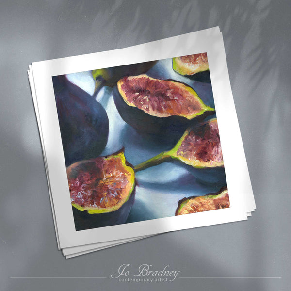 A stack of square art prints on archival paper on a gray kitchen counter. The prints show fresh purple and pink figs, scattered on a blue background. This is a giclee print of my realistic oil painting fruit still life. The original artwork is sold.
