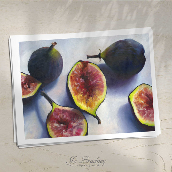 A stack of horizontal art prints on archival paper on a wood kitchen counter. The prints show fresh cut Mission figs on a pale blue background. This is a giclee print of my realistic oil painting fruit still life. The original artwork is sold.