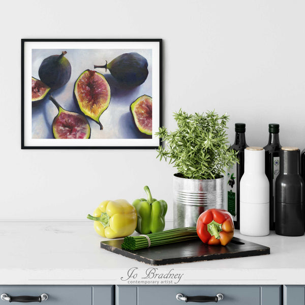 A fresh fig painting print in a simple, elegant black picture frame on a kitchen wall. There is a chopping board, vegetables, herbs and some bottles. The art print is as large as three bell peppers. The largest size is 11 inches high by 14 wide