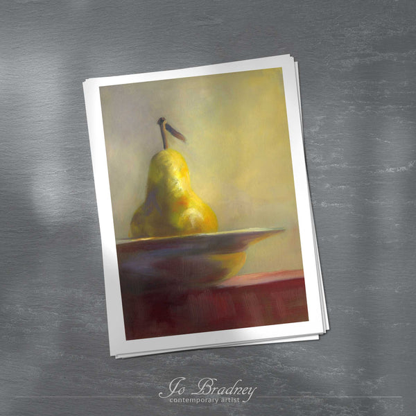 A stack of vertical art prints on archival paper on a wood kitchen counter. The prints show a gold fall pear in a china bowl. This is a giclee print of my realistic oil painting still life. The original artwork is sold.